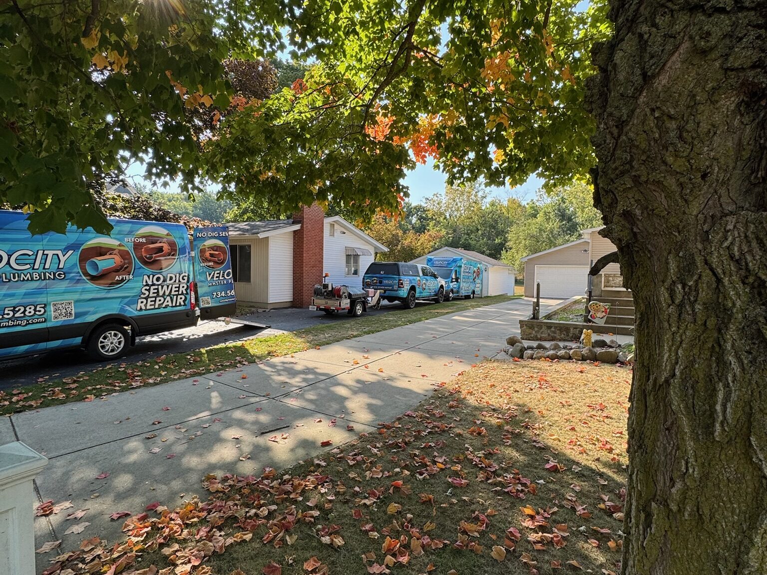 Two Velocity Master Plumbing service trucks parked curbside under autumn trees during a sewer camera inspection visit at an Ann Arbor home
